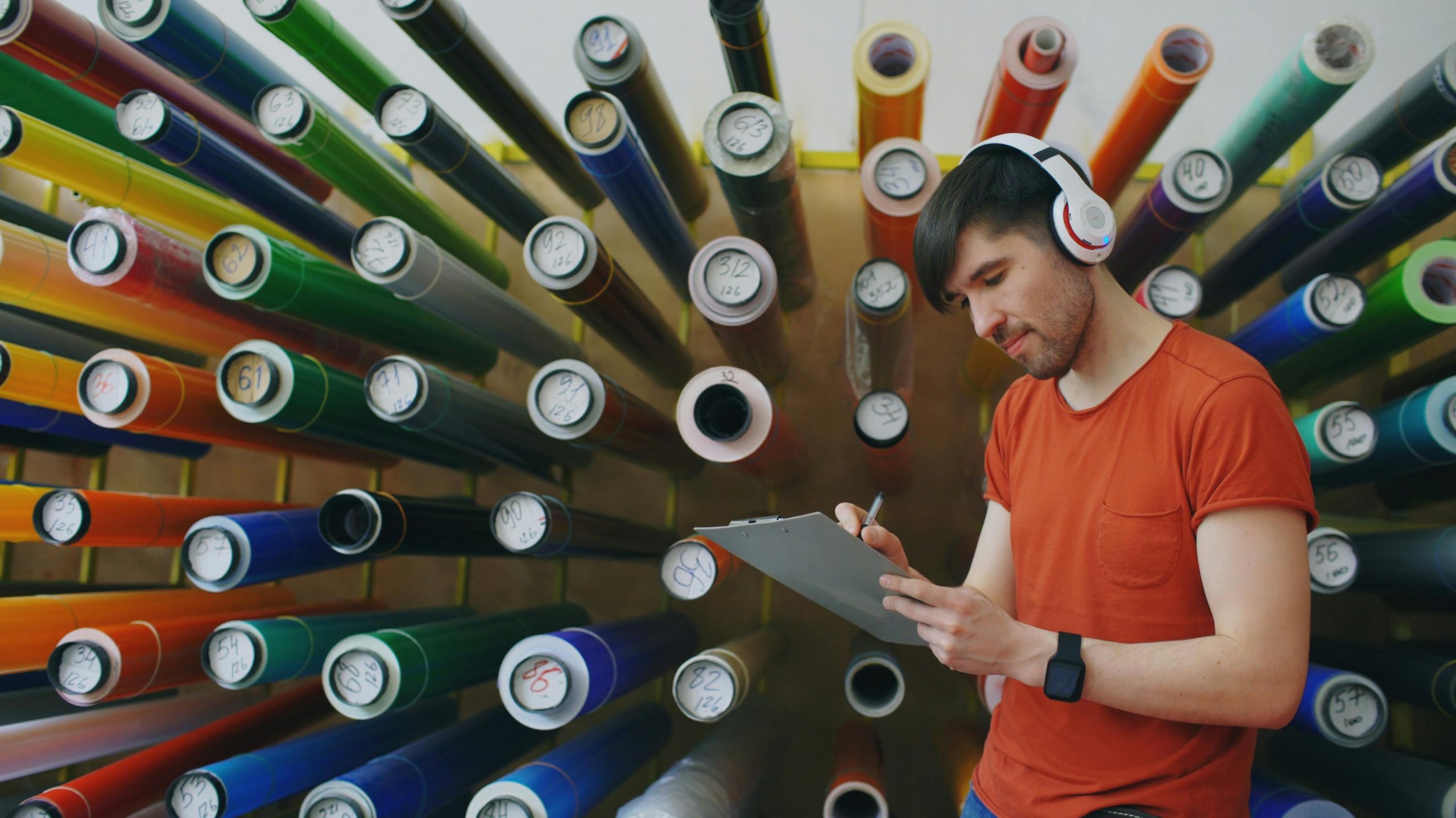 Man in a design studio with colorful rolls and headphones, noting creative ideas.